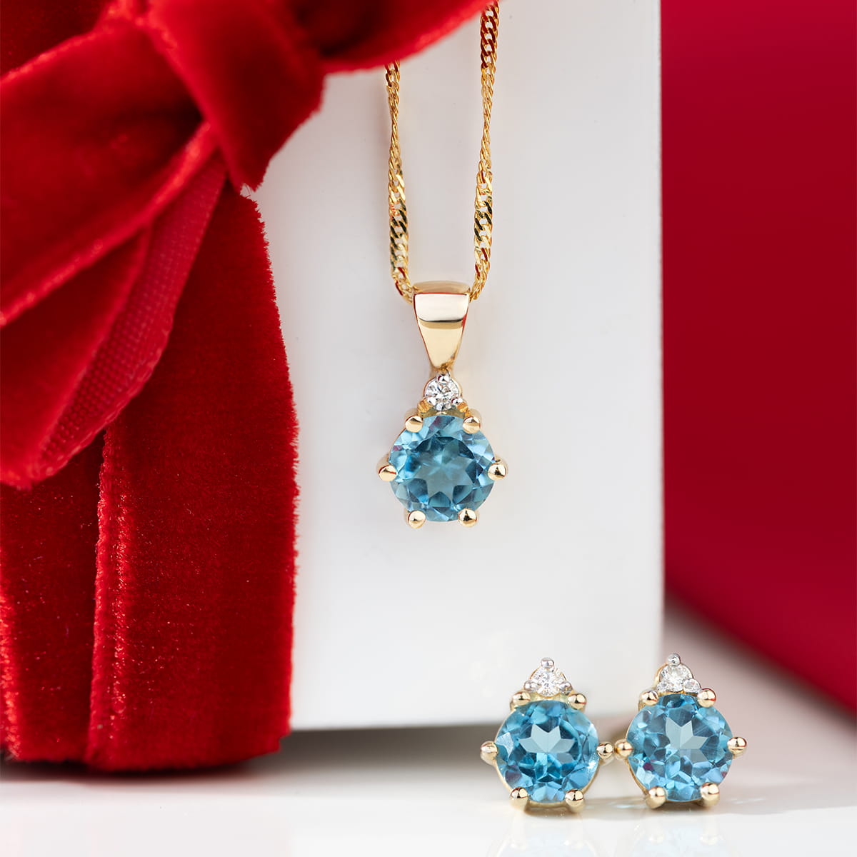 K0076.Z3.TOB.01_4 Złoty naszyjnik z niebieskim kamieniem i kolczyki w tym samym stylu, na tle czerwonego materiału.