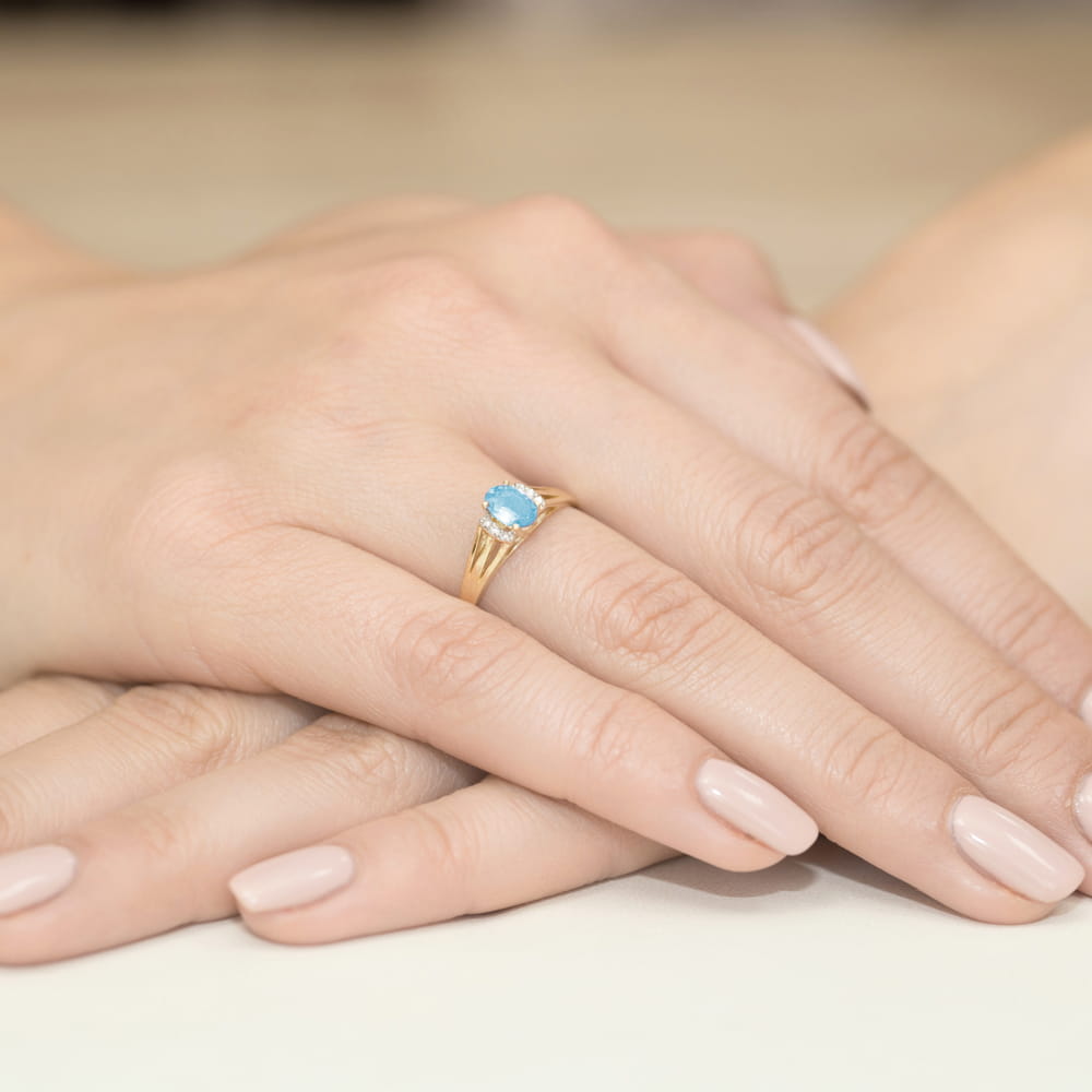 W0005.Z3.TOB.01_4 Złoty pierścionek z niebieskim kamieniem na palcu, dłonie z delikatnym manicure w jasnym kolorze.