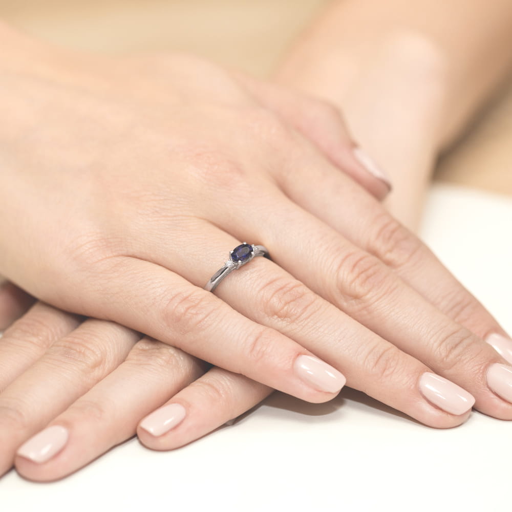 W0006.B3.SAB.01_4 Na dłoni widoczny pierścionek z niebieskim kamieniem, otoczony delikatnymi zdobieniami.