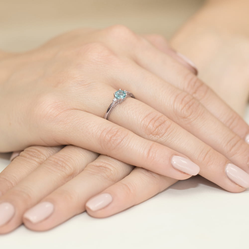 W0010.B3.TOB.01_4 Dwie dłonie z delikatnym pierścionkiem z niebieskim kamieniem na palcu. Paznokcie pomalowane na jasny kolor.