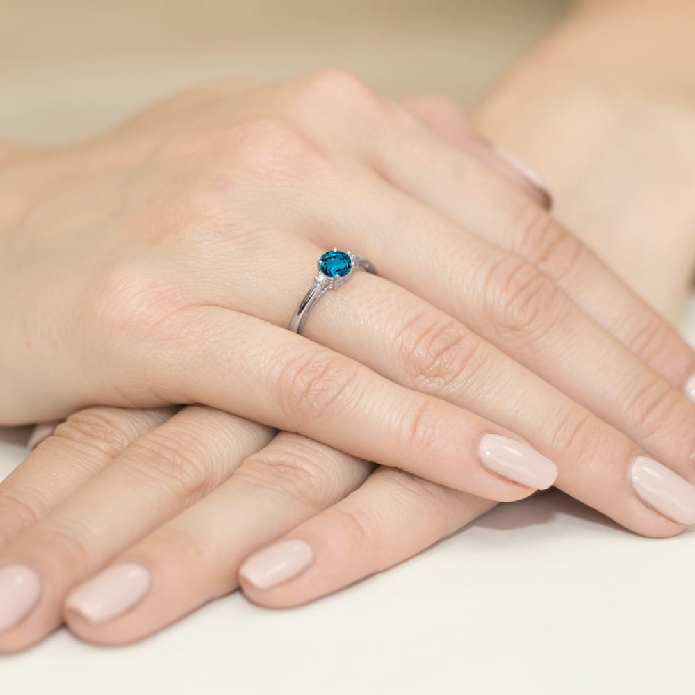 W0010.B3.TOL.01_4 Na dłoni z delikatnym manicure znajduje się srebrny pierścionek z niebieskim kamieniem.