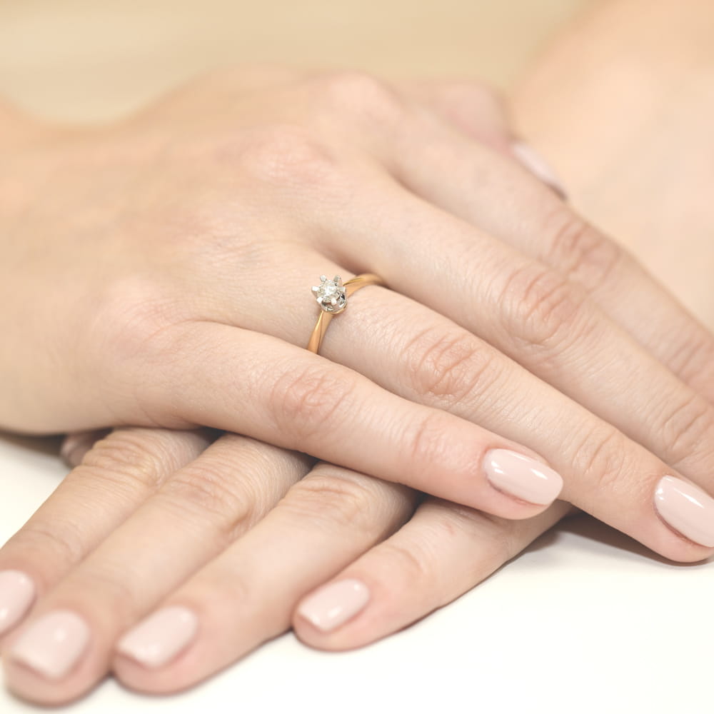 W0036.Z3.DIW.03_4 Złoty pierścionek z diamentem na palcu jednej ręki, trzymanej na dłoniach drugiej osoby.