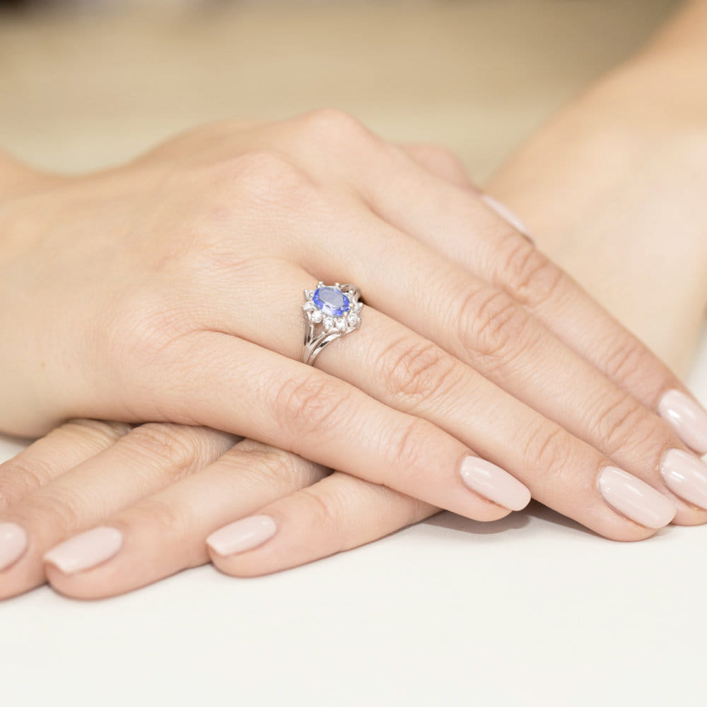 W0037.B3.TAP.01_4 Na dłoni z delikatnym manicure znajduje się srebrny pierścionek z niebieskim kamieniem i diamentami.