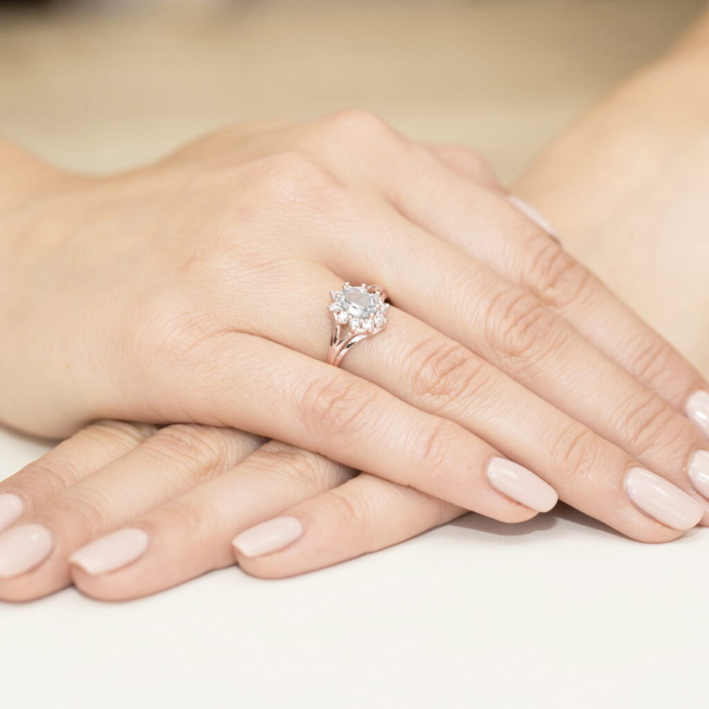 Na dłoni z delikatnym manicure znajduje się pierścionek z dużym, błyszczącym kamieniem.