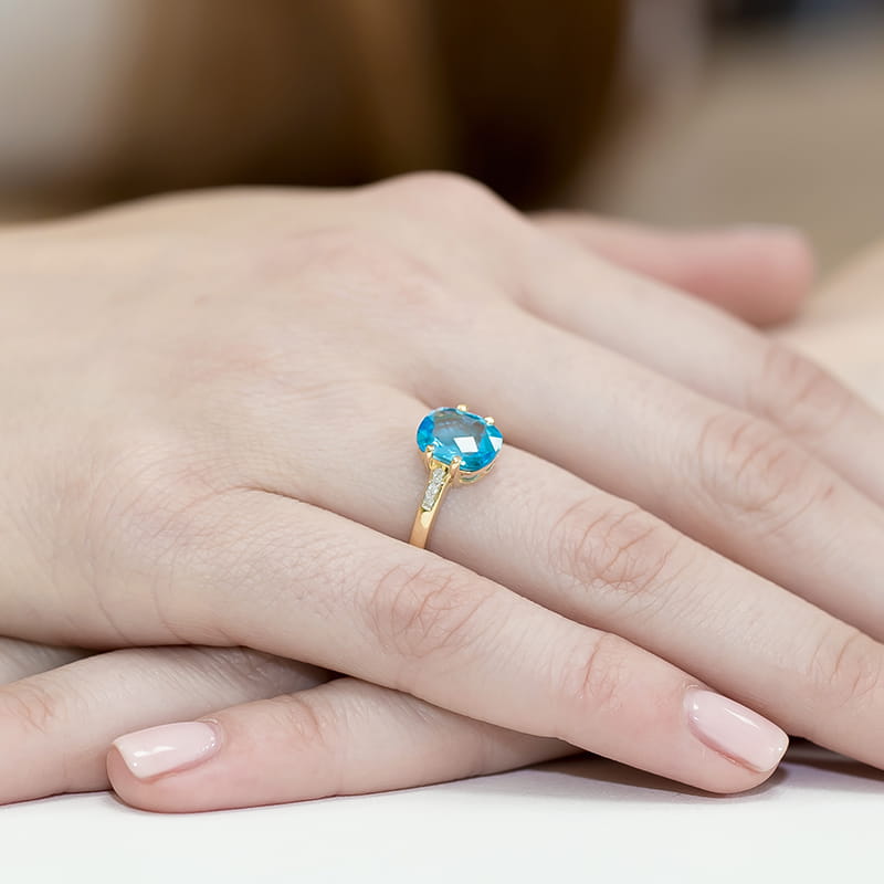 W0038.Z3.TOB.01_4 Złoty pierścionek z dużym, niebieskim kamieniem i drobnymi diamentami na obrączce, na dłoni z zadbanymi paznokciami.