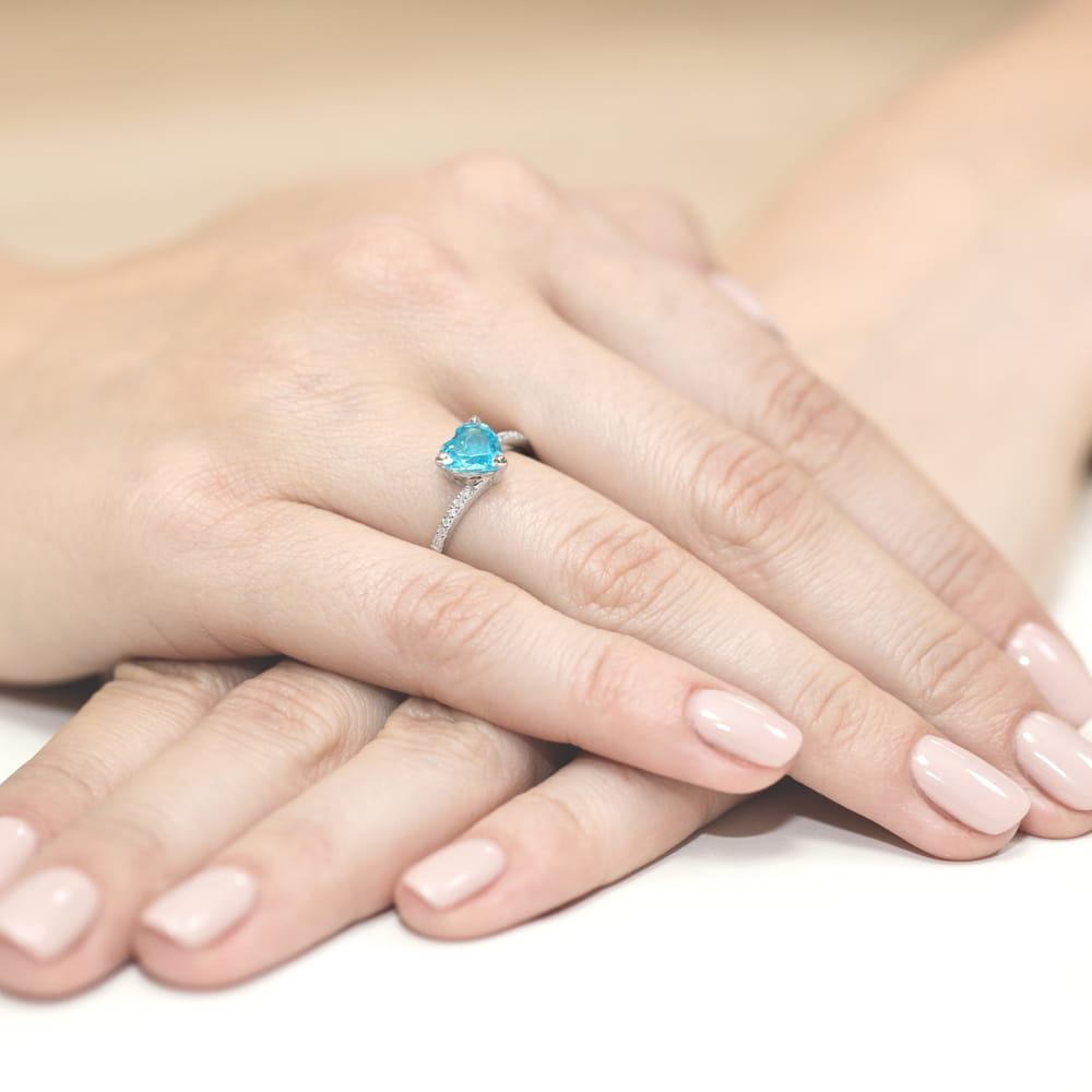 W0050.B3.TOB.01_4 Srebrny pierścionek z sercowym, niebieskim kamieniem na palcu, otoczony dłońmi z delikatnym manicure.