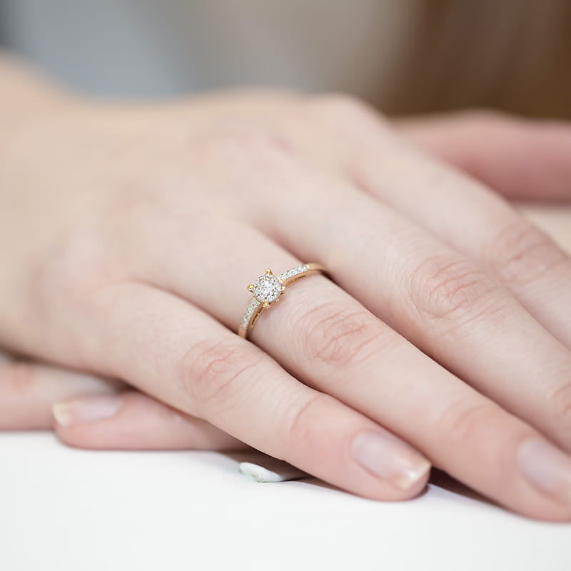 W0055.Z3.DIW.01_4 Złoty pierścionek z diamentem na palcu kobiecej ręki, delikatne dłonie na jasnym tle.