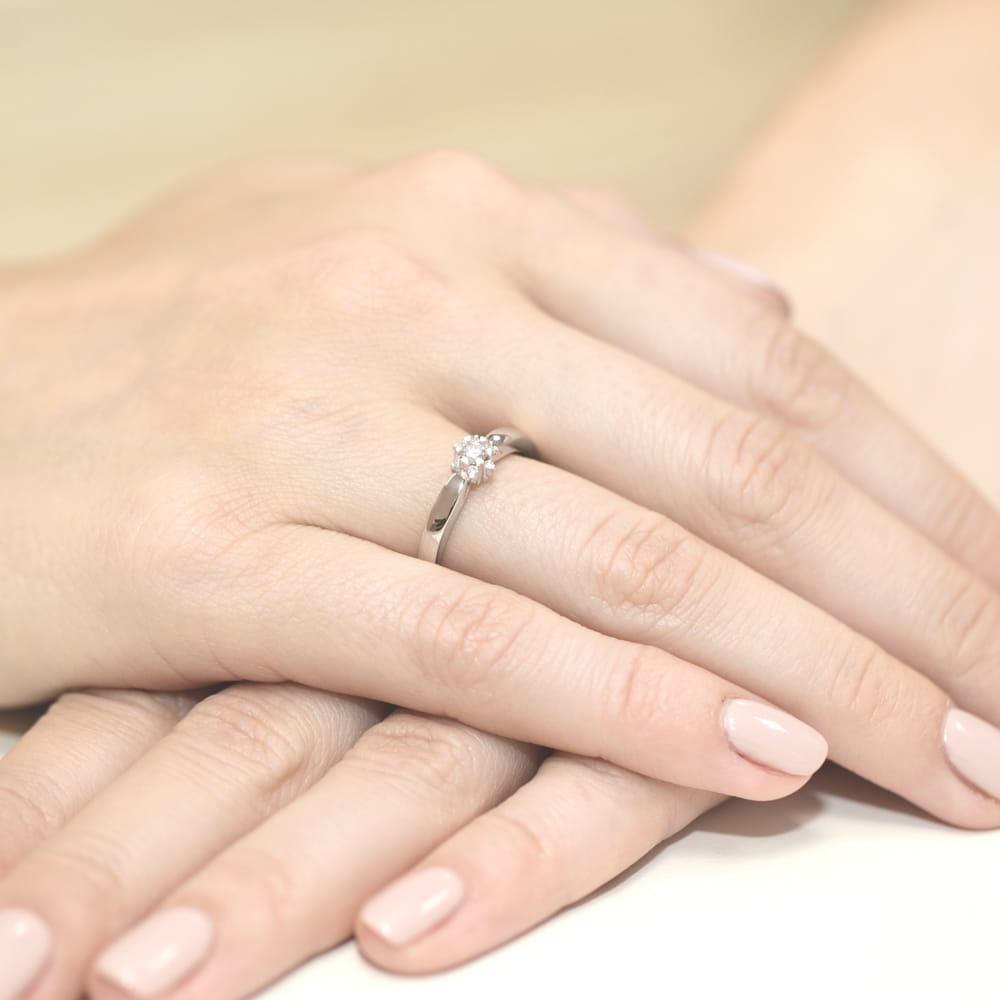 W0061.B3.DIW.01_4 Na dłoni znajduje się srebrny pierścionek z centralnym diamentem, otoczony mniejszymi kamieniami.