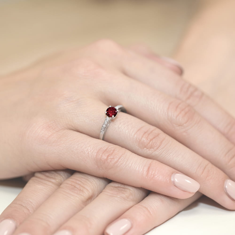 W0066.B3.GAR.01_4 Pierścionek z czerwonym kamieniem na palcu, otoczony drobnymi diamentami, na tle dłoni z zadbanymi paznokciami.