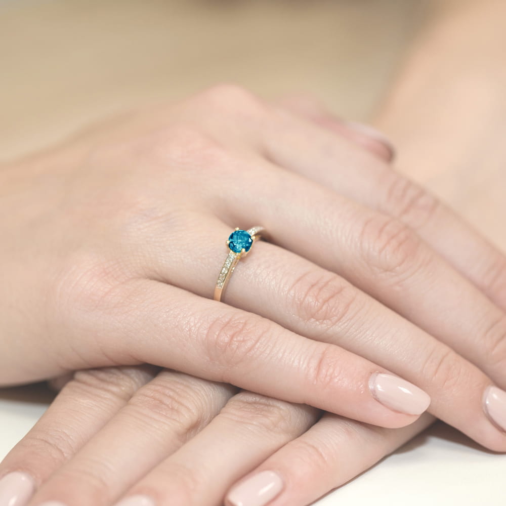 W0066.Z3.TOL.01_4 Złoty pierścionek z niebieskim kamieniem i drobnymi diamentami na delikatnej dłoni.