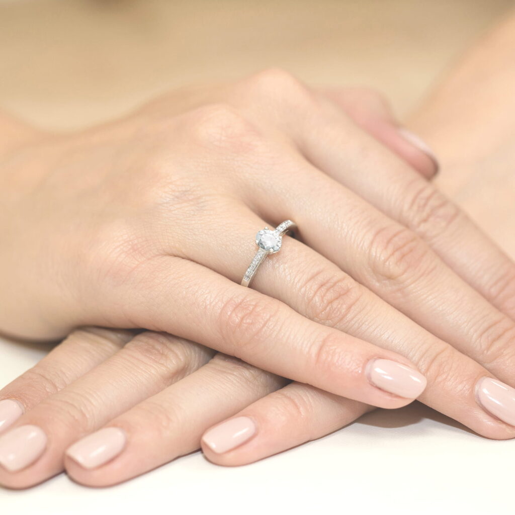 Na dłoni z delikatnym manicure znajduje się pierścionek z dużym, błyszczącym kamieniem.
