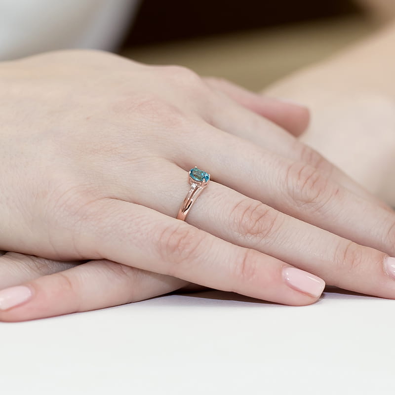 W0068.R3.TOB.01_4 Ręka z delikatnym pierścionkiem z niebieskim kamieniem na tle jasnego stołu.