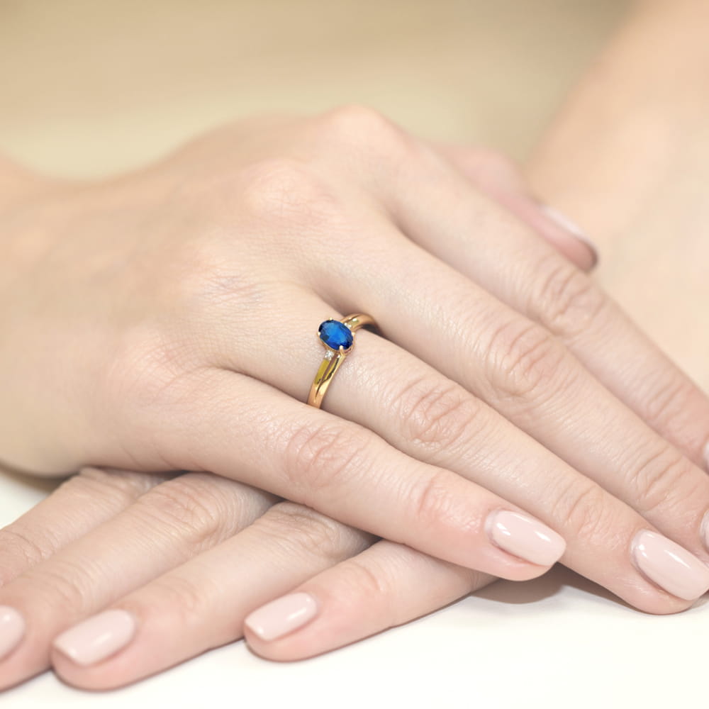 W0068.Z3.SAB.01_4 Złoty pierścionek z niebieskim kamieniem na palcu, dłonie z delikatnym manicure.