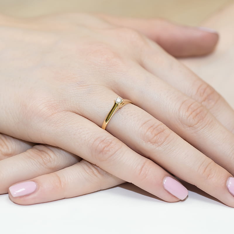 W0070.Z3.DIW.01_4 Złoty pierścionek z małym diamentem na palcu kobiecej ręki z różowym manicure.