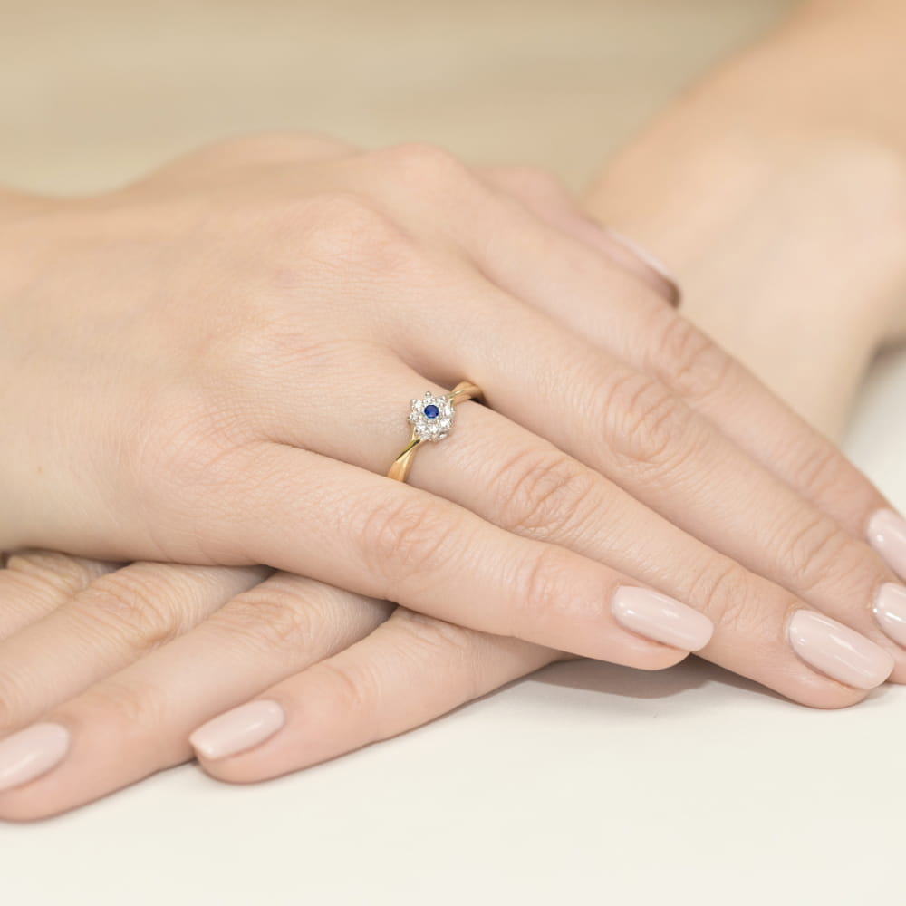 W0072.Z3.SAB.01_4 Złoty pierścionek z niebieskim kamieniem i diamentami na dłoni z zadbanymi paznokciami.