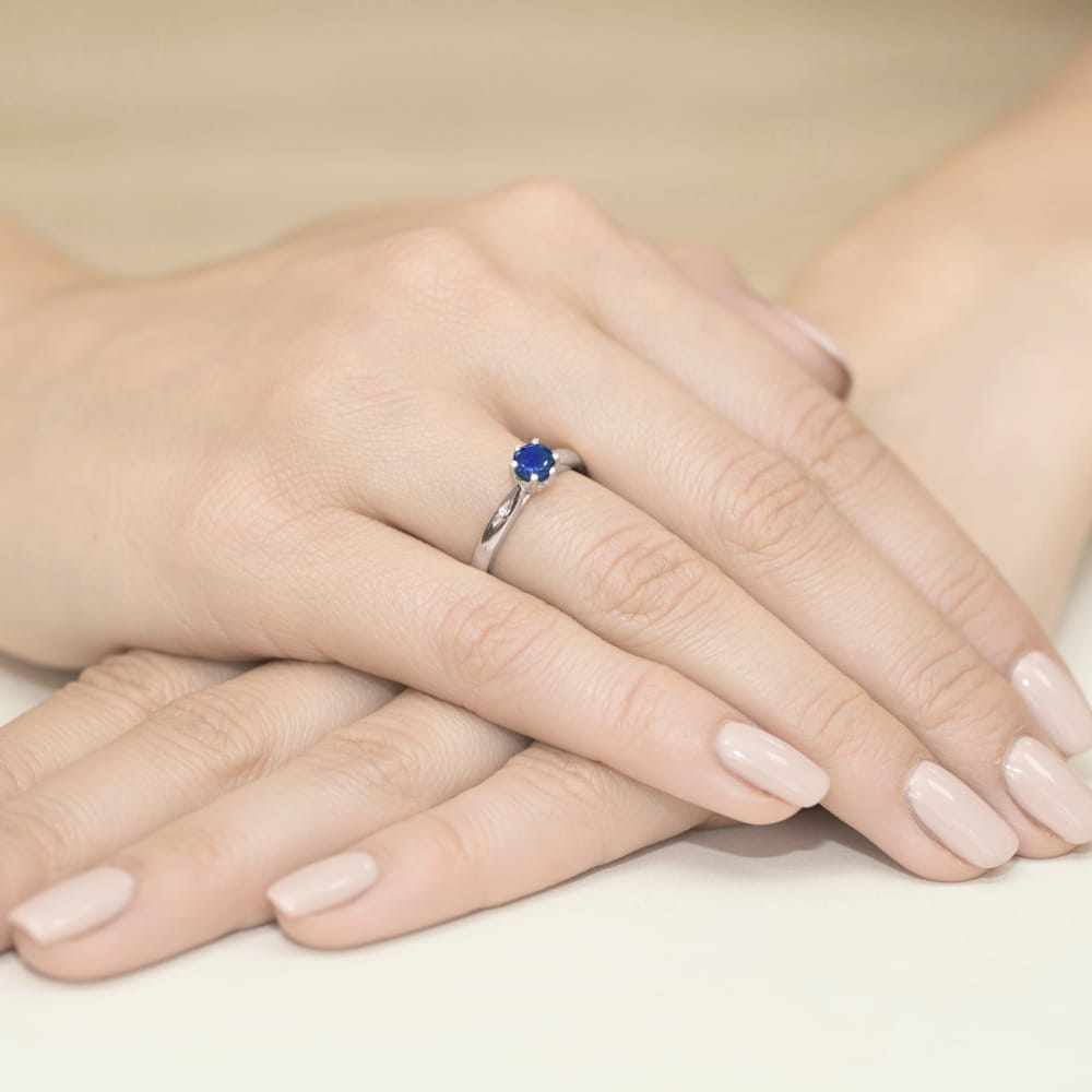 W0074.B3.SAB.01_4 Srebrny pierścionek z niebieskim kamieniem na palcu, dłonie z delikatnym manicure.