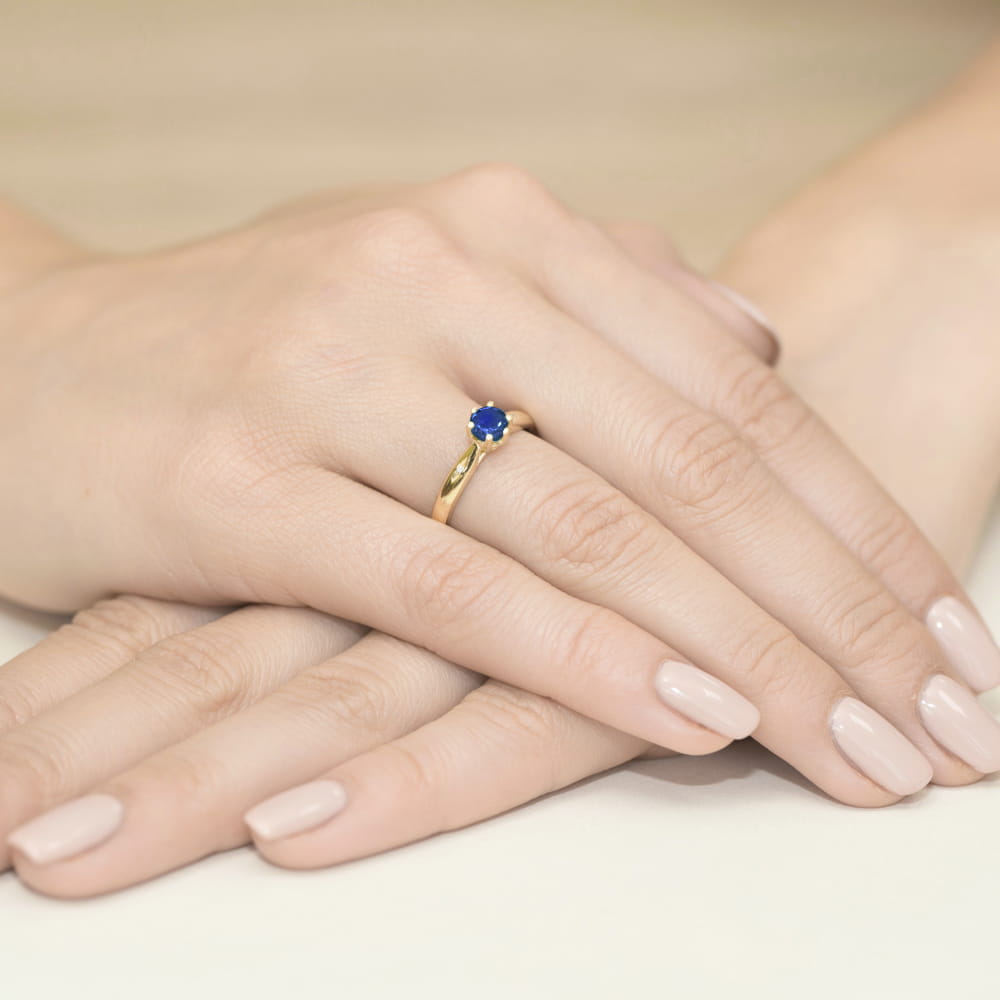 W0074.Z3.SAB.01_4 Złoty pierścionek z niebieskim kamieniem na palcu, dłonie z delikatnym manicure.