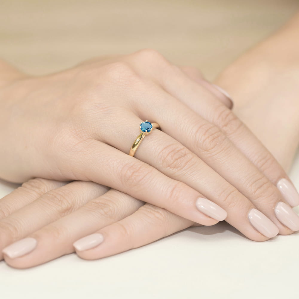 W0074.Z3.TOL.01_4 Złoty pierścionek z niebieskim kamieniem na palcu, dłonie z delikatnym manicure.