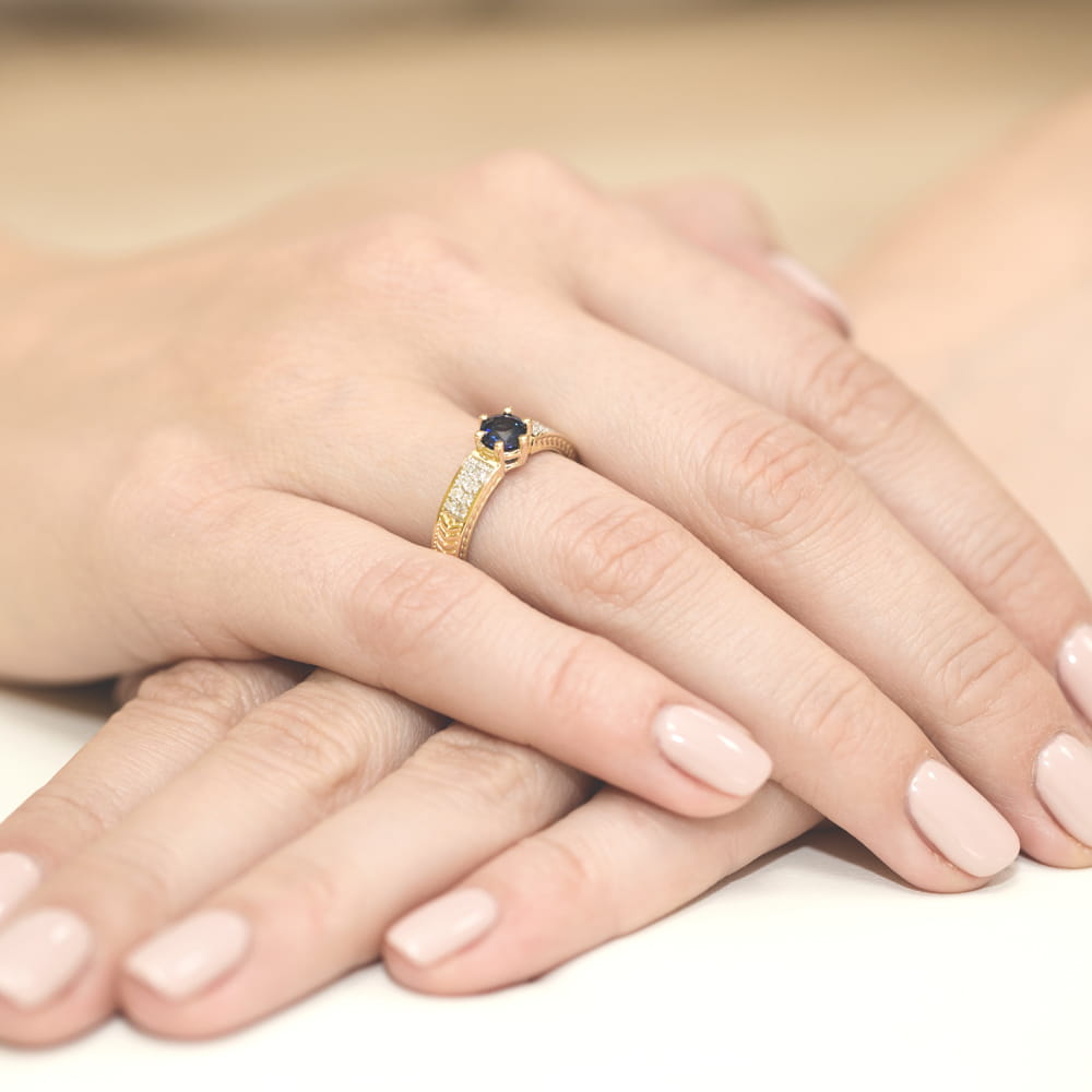W0075.Z3.SAB.01_4 Złoty pierścionek z niebieskim kamieniem i diamentami na tle dłoni z delikatnym manicure.