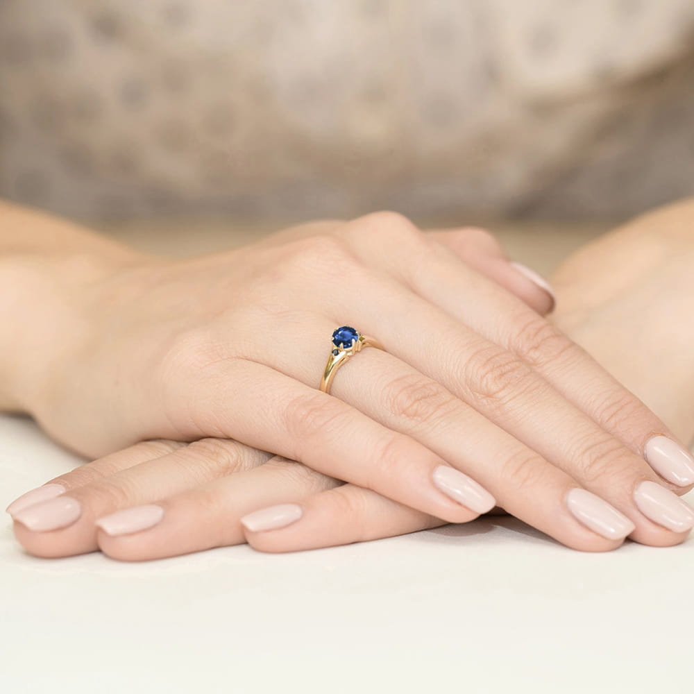 W0076.Z3.SAB.02_3 Złoty pierścionek z niebieskim kamieniem na palcu, dłonie z delikatnym manicure w jasnym kolorze.