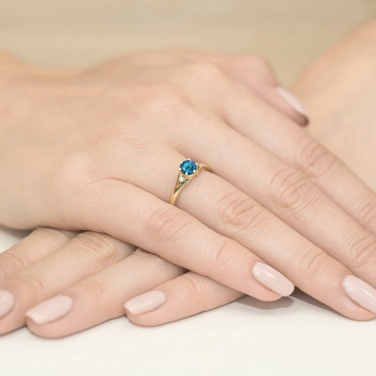 W0076.Z3.TOL.01_4 Złoty pierścionek z niebieskim kamieniem na palcu, dłonie z delikatnym manicure w jasnym kolorze.