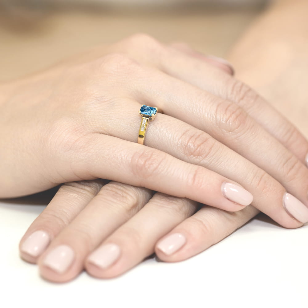 W0078.Z3.TOB.01_4 Złoty pierścionek z niebieskim kamieniem na palcu, dłonie z delikatnym manicure w jasnym kolorze.