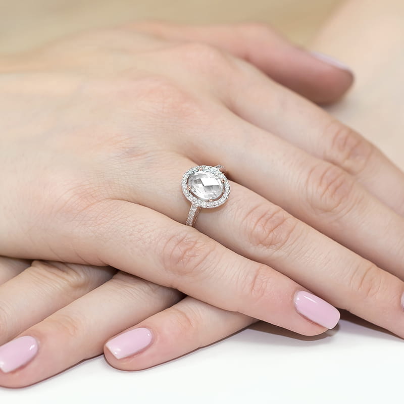 W0080.R3.LDW.01_4 Pierścionek z dużym, przezroczystym kamieniem otoczonym małymi diamentami na dłoni z różowym manicure.