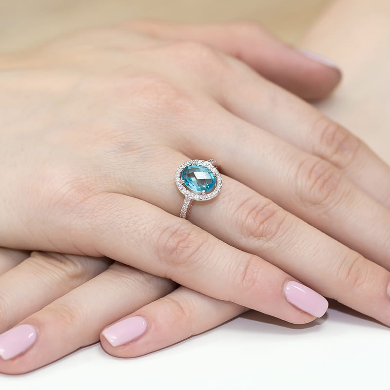W0080.R3.TOB.01_4 Pierścionek z niebieskim kamieniem otoczony diamentami na dłoni z różowym manicure.