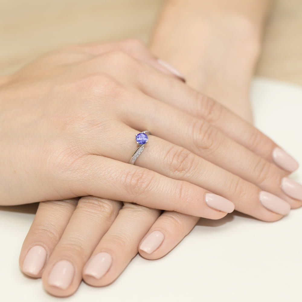 W0084.B3.TAP.01_4 Dwie ręce z delikatnym manicure, jedna z pierścionkiem z fioletowym kamieniem i diamentowymi wstawkami.