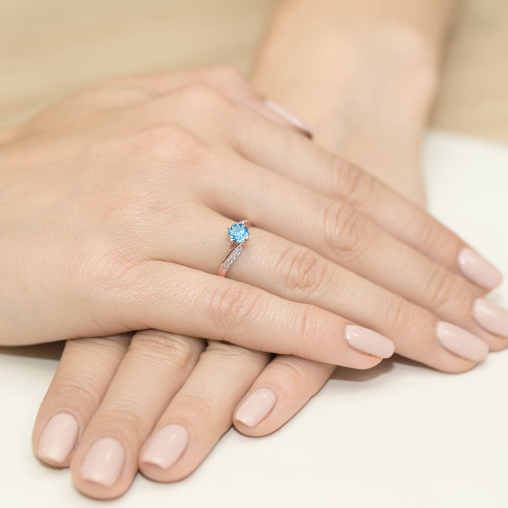 W0084.R3.TOB.01_4 Na dłoni z delikatnym manicure znajduje się pierścionek z niebieskim kamieniem i drobnymi diamentami.