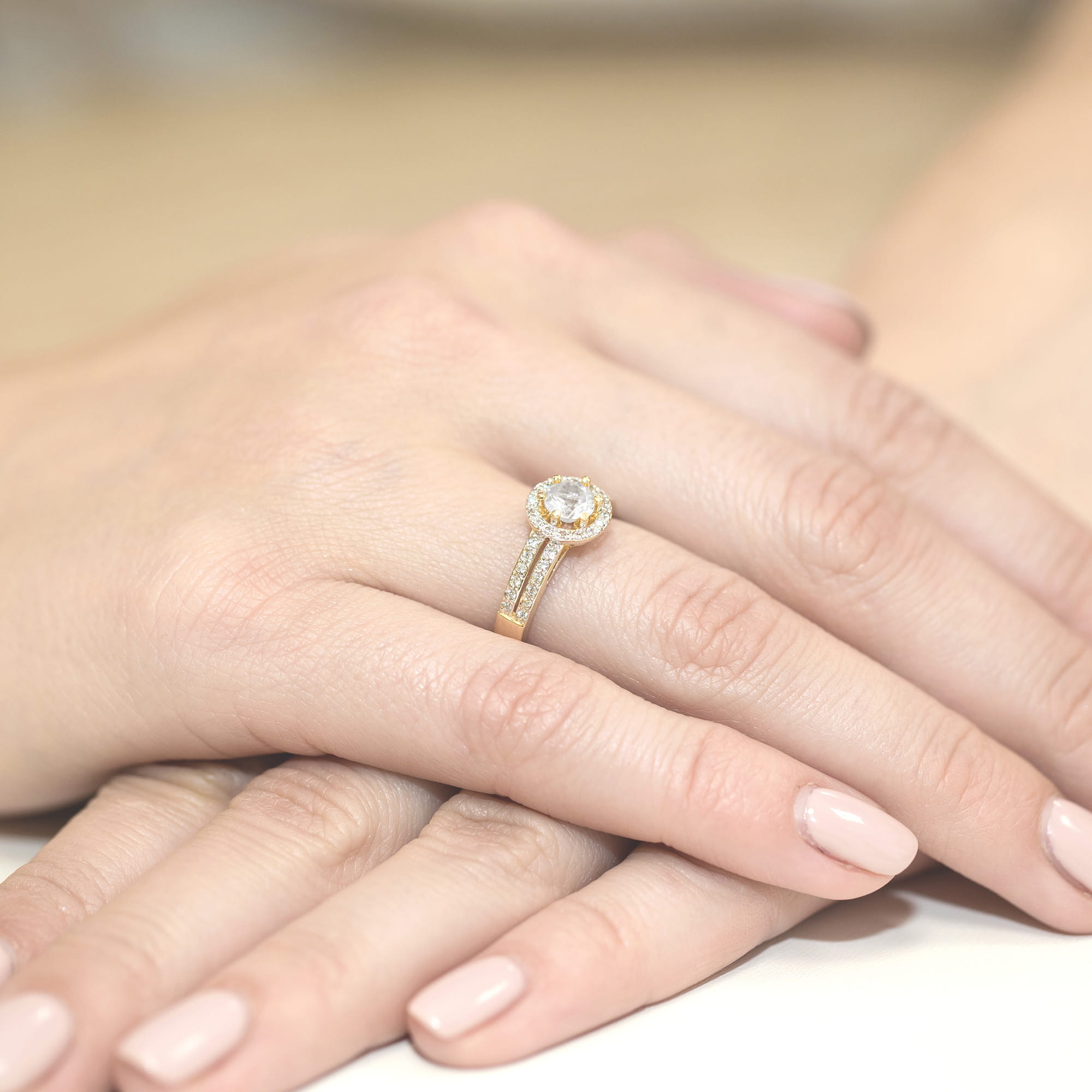 W0087.Z3.LDW.01_4 Złoty pierścionek z okrągłym diamentem i drobnymi kamieniami, na dłoni z delikatnym manicure.