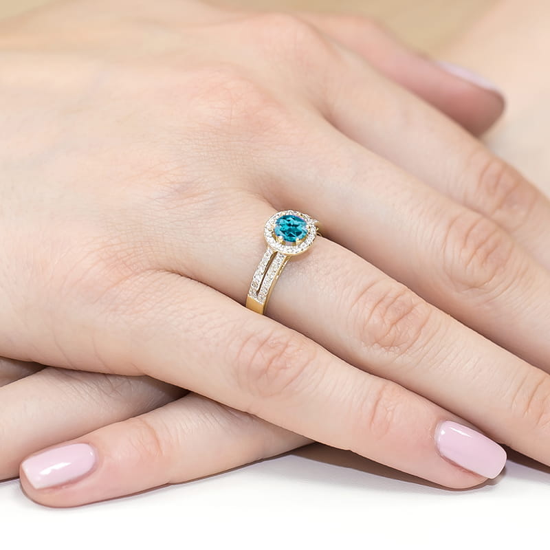 W0087.Z3.TOL.01_4 Złoty pierścionek z niebieskim kamieniem i diamentowymi zdobieniami na palcu z różowym manicure.