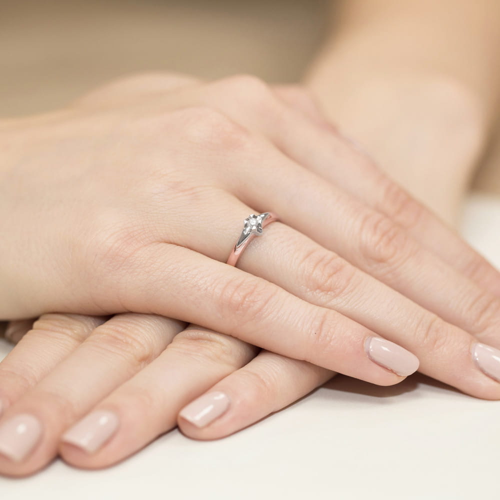 W0088.R3.DIW.02_4 Na dłoni z delikatnym manicure znajduje się pierścionek z diamentem, otoczony subtelnymi zdobieniami.