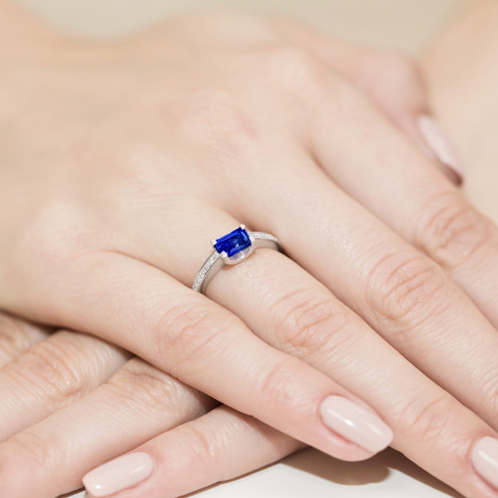 W0105.B3.SAB.01_4 Pierścionek z niebieskim kamieniem szlachetnym na palcu, otoczony delikatnymi, srebrnymi zdobieniami.