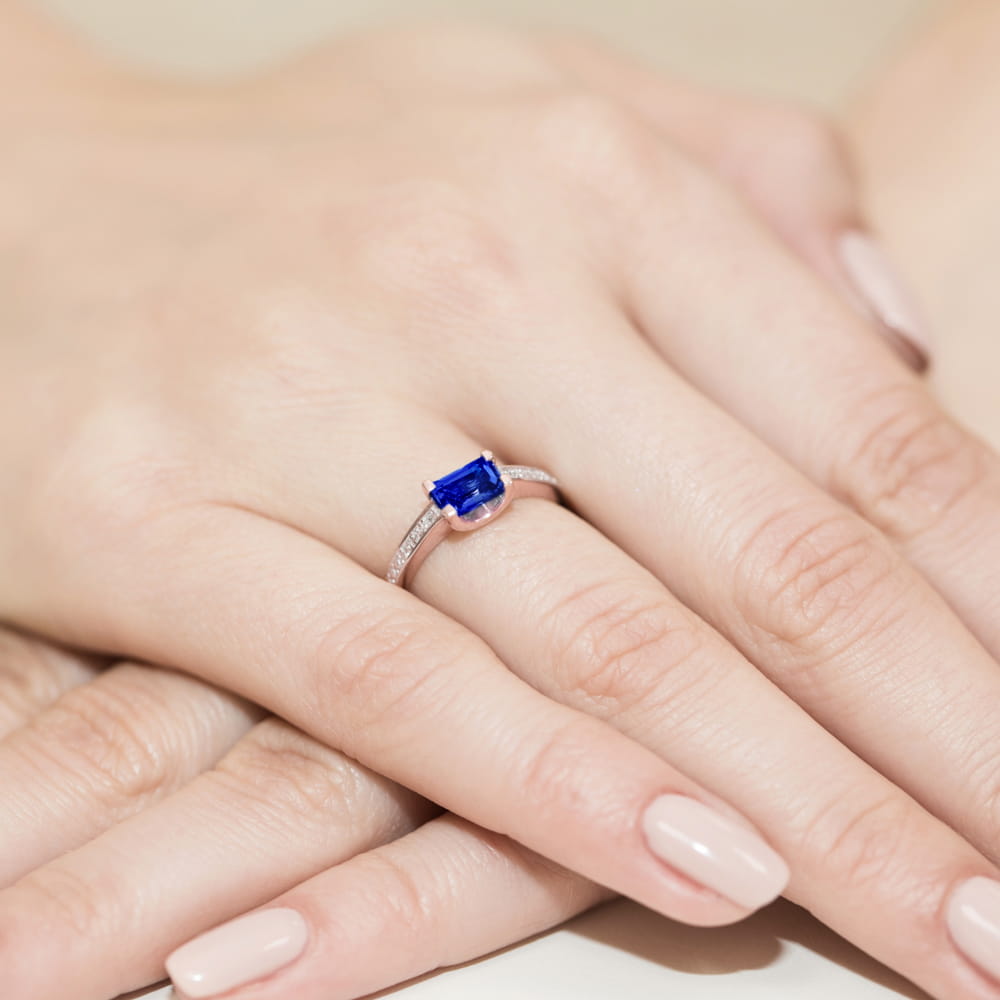 W0105.R3.SAB.01_4 Pierścionek z niebieskim kamieniem szlachetnym na palcu, otoczony delikatnymi, błyszczącymi diamentami.