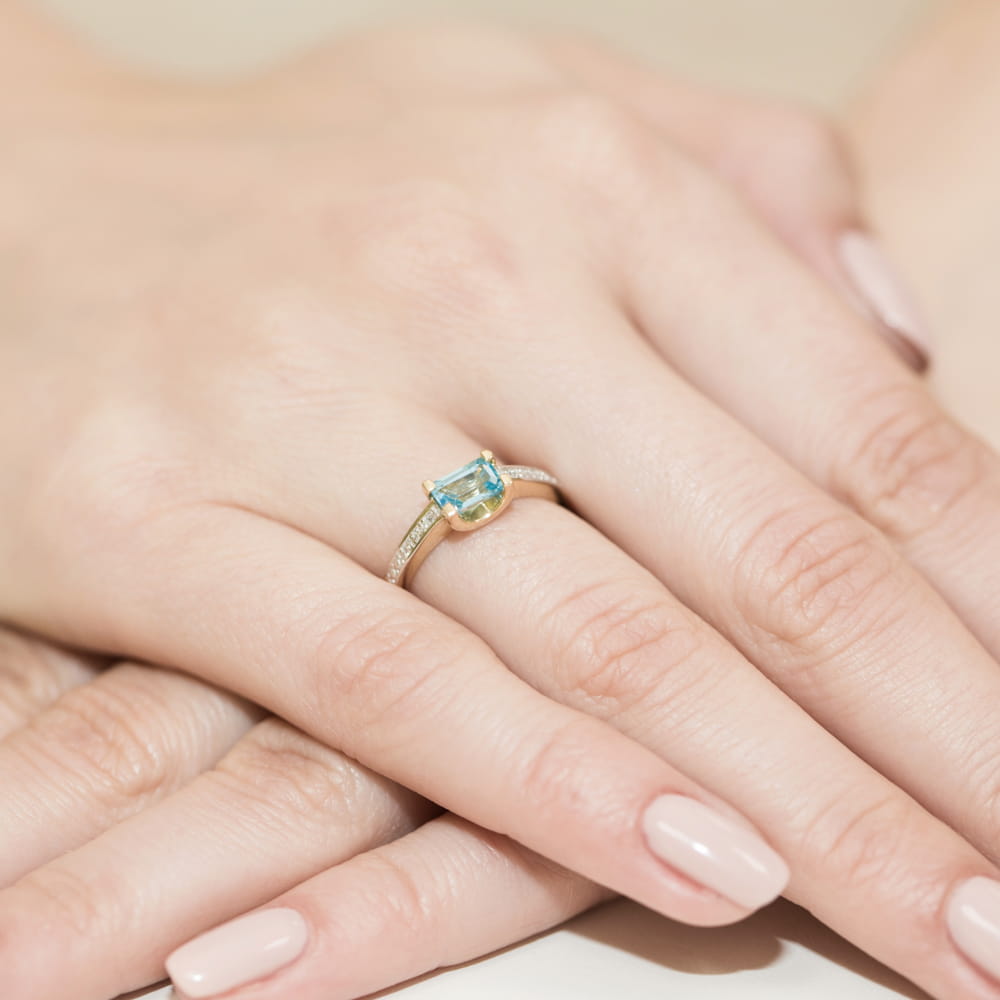 W0105.Z3.TOB.01_4 Pierścionek z niebieskim kamieniem na palcu, otoczony delikatnymi, złotymi zdobieniami.