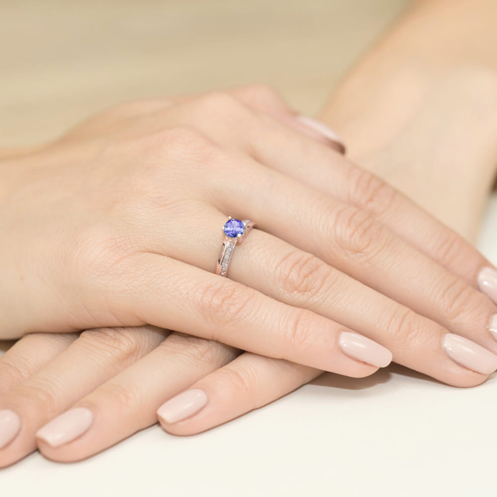 W0106.R3.TAP.01_4 Na dłoni z delikatnym manicure znajduje się pierścionek z niebieskim kamieniem i ozdobnymi detalami.