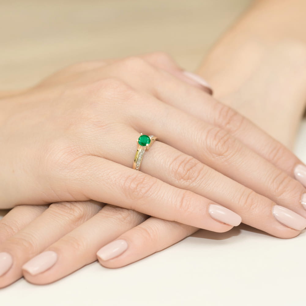 W0106.Z3.EMG.01_4 Złoty pierścionek z zielonym kamieniem na dłoniach z delikatnym manicure w odcieniu nude.