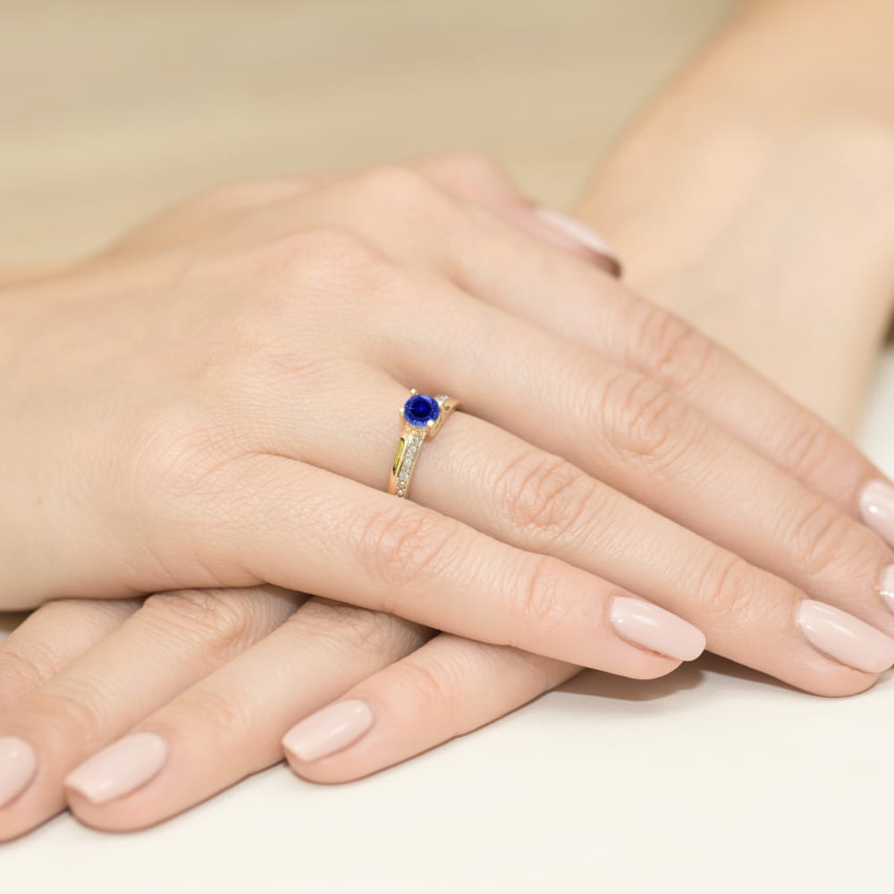 W0106.Z3.SAB.01_4 Złoty pierścionek z niebieskim kamieniem na palcu, dłonie z delikatnym manicure w jasnym kolorze.