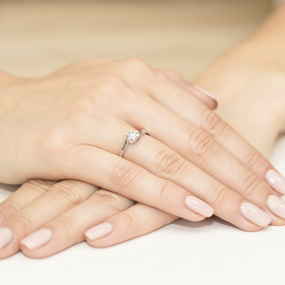 W0163.B3.DIW.03_4 Dwie dłonie z delikatnym pierścionkiem z diamentem na palcu, z zadbanymi, jasnymi paznokciami.