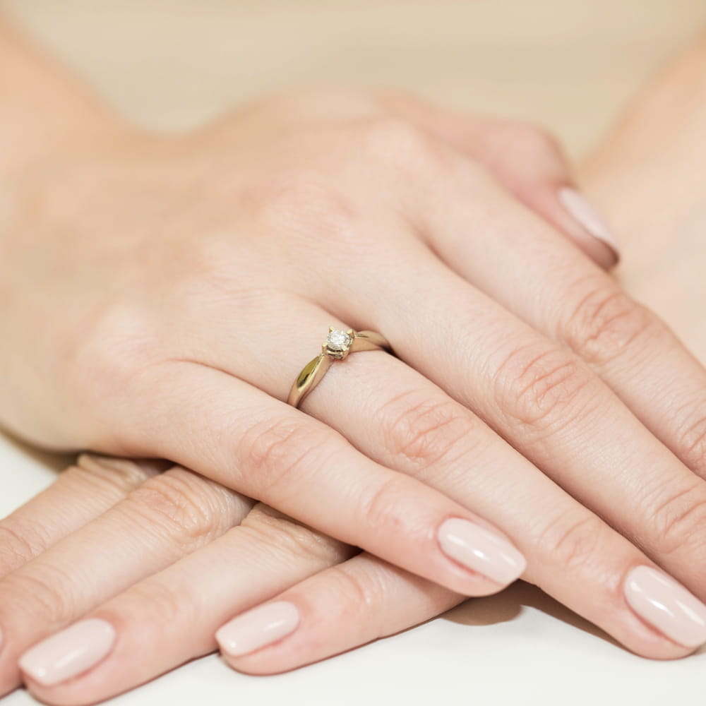 W0171.Z3.DIW.01_4 Złoty pierścionek z diamentem na palcu kobiety z zadbanymi, jasnymi paznokciami.
