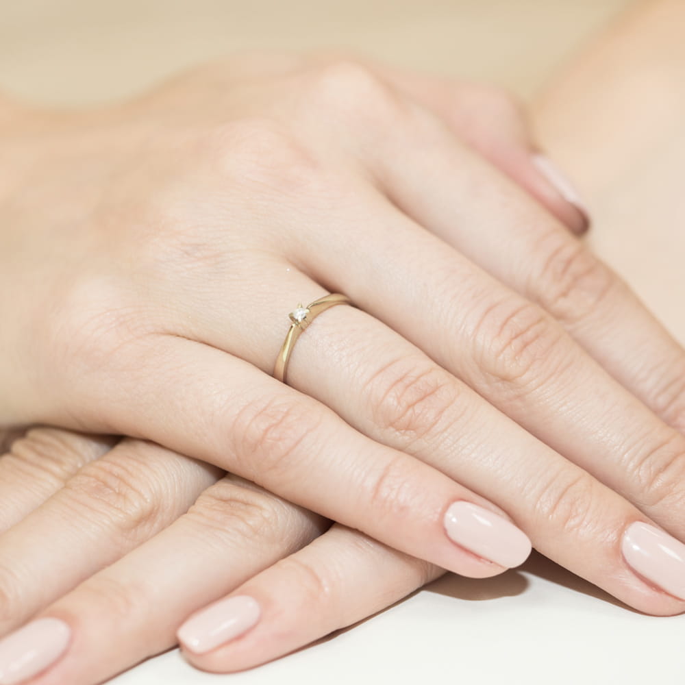 W0175.Z3.DIW.01_4 Złoty pierścionek z małym diamentem na palcu kobiety, obok dłonie innej osoby.