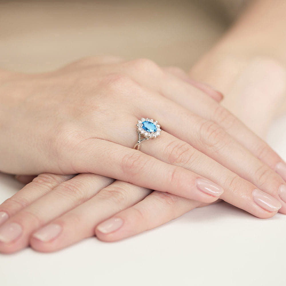 W0182.B3.TOB.01_4 Na dłoni widoczny pierścionek z niebieskim kamieniem w otoczeniu drobnych, jasnych diamentów.