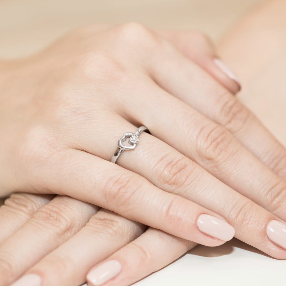 W0188.B3.DIW.01_4 Srebrny pierścionek w kształcie serca z małym diamentem na palcu z zadbanymi, różowymi paznokciami.