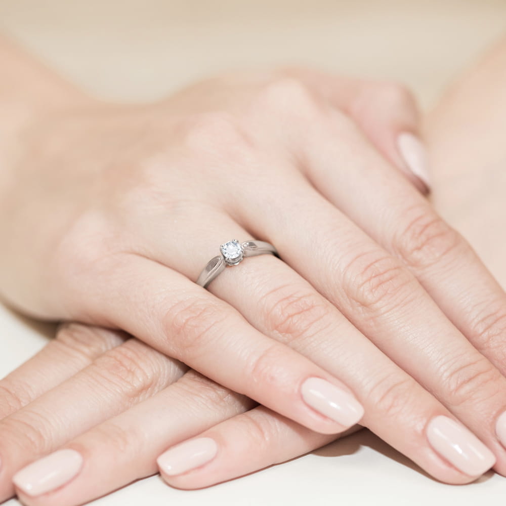 W0197.B3.DIW.01_4 Na dłoni znajduje się srebrny pierścionek z centralnym diamentem, otoczony delikatnymi palcami.