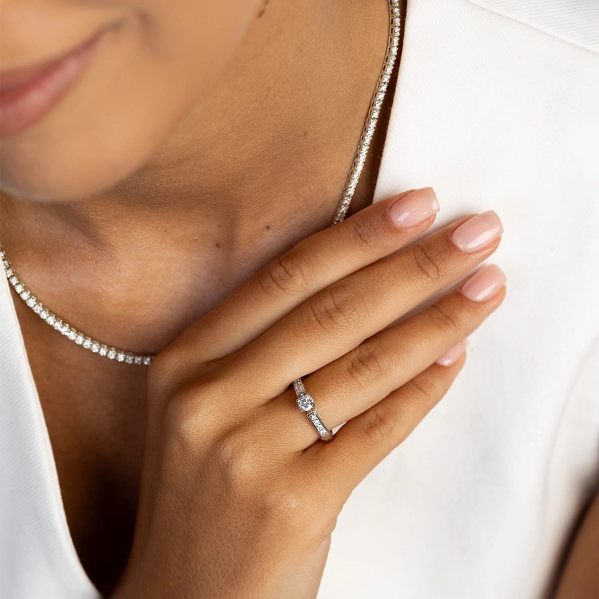 W0198.B3.DIW.01_4 Kobieta z delikatnym naszyjnikiem i pierścionkiem z diamentem na palcu, trzyma rękę przy twarzy.