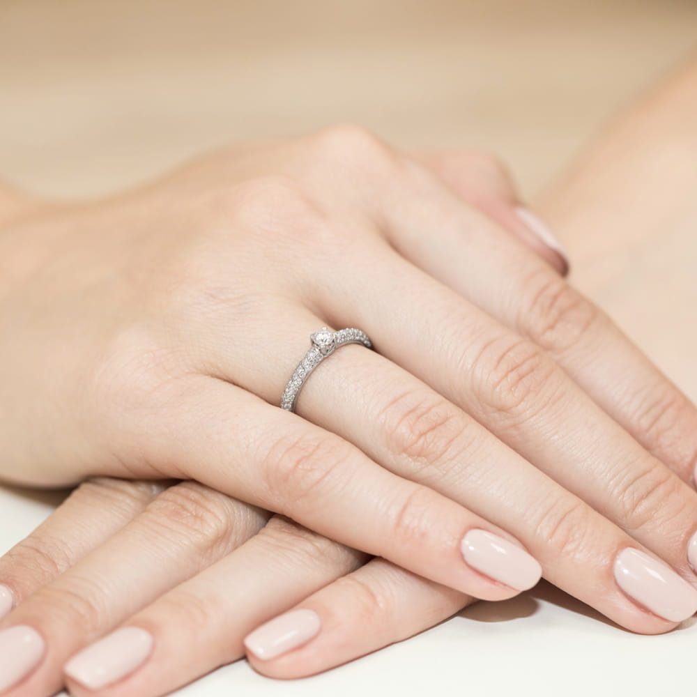 W0200.B3.DIW.01_4 Na dłoni widoczny jest srebrny pierścionek z diamentem, otoczony drobnymi kamieniami.