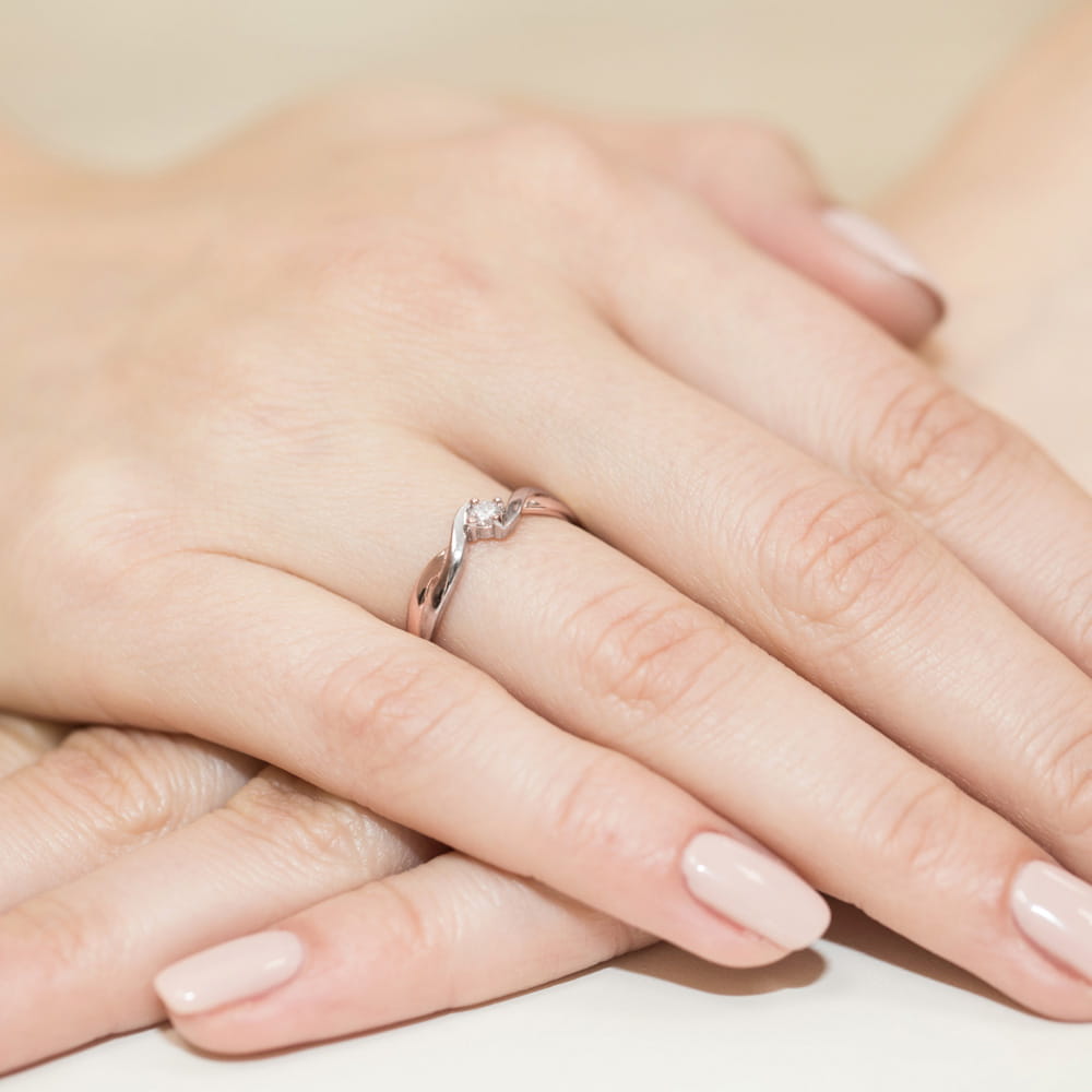 W0208.R3.DIW.01_4 Na dłoni widoczny srebrny pierścionek z delikatnym, skręconym wzorem i małym diamentem.