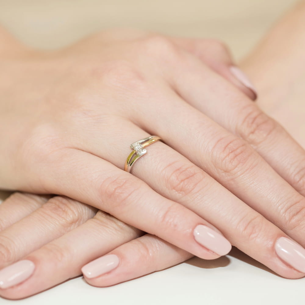 W0210.Z3.DIW.02_4 Dwie ręce z delikatnym złotym pierścionkiem z diamentem na palcu. Paznokcie w jasnym kolorze.