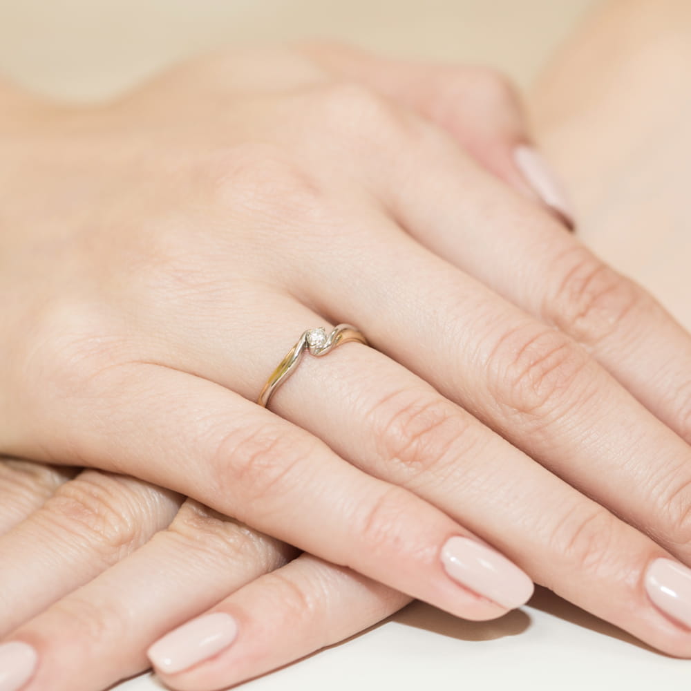 W0211.Z3.DIW.01_4 Dwie dłonie z delikatnym pierścionkiem na palcu, z małym diamentem, na tle jasnej powierzchni.